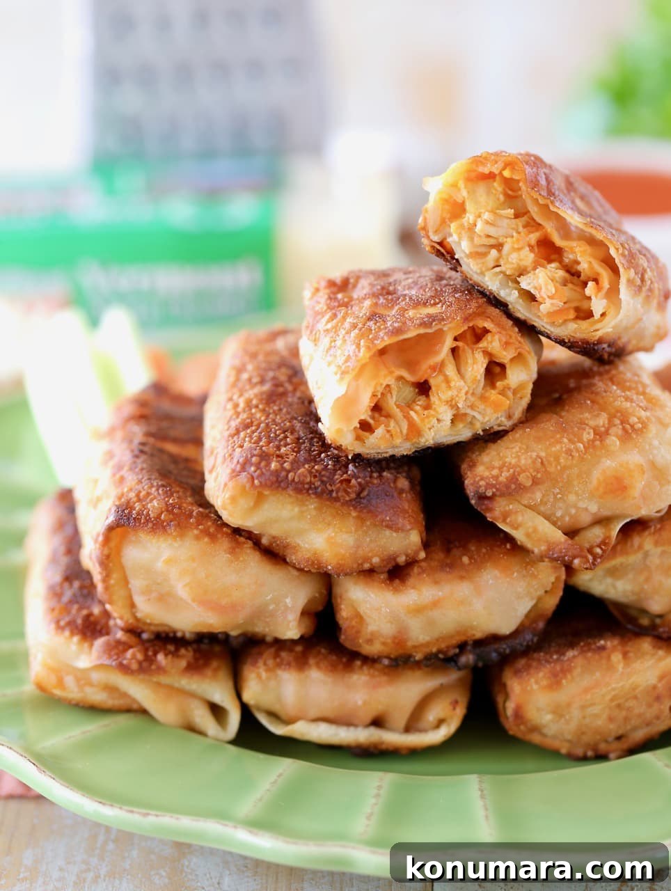 Crispy Buffalo Chicken Egg Rolls Recipe with Blue Cheese Dip 2 Buffalo chicken egg rolls piled up on a green plate with one egg roll cut in half on top, with cheddar cheese and a cheese grater in the background