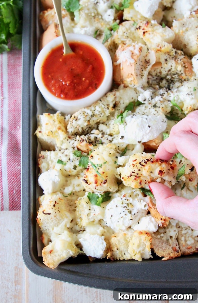 Cheesy Garlic Bread Nachos Recipe 5 Hand grabbing a piece of garlic bread nachos off of a baking sheet with a small bowl of marinara sauce in the corner