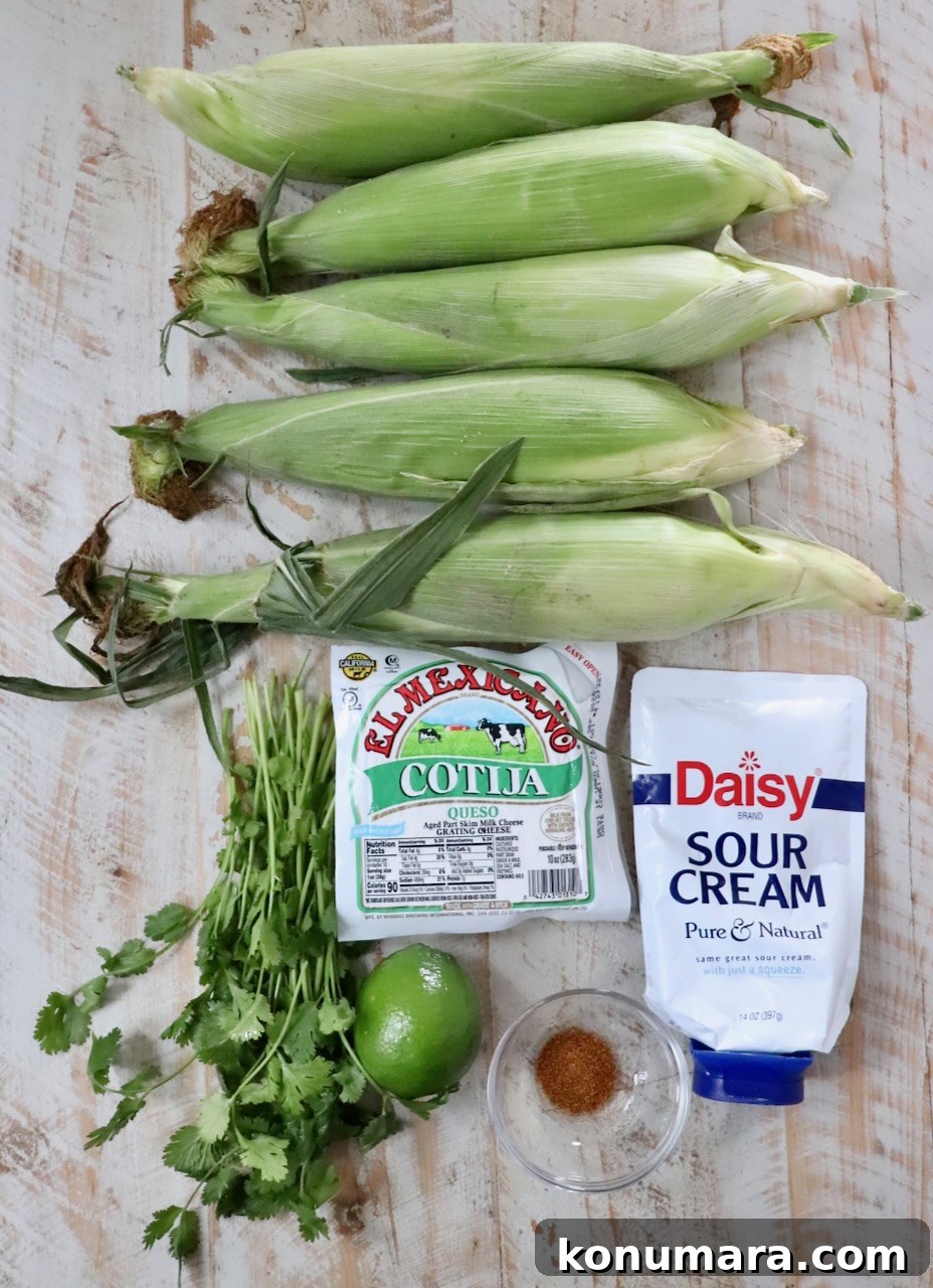 5 ears of corn on the cob, next to a package of cotija cheese, sour cream, chili powder in a small bowl, a lime, and cilantro, all lined up on a white wooden board