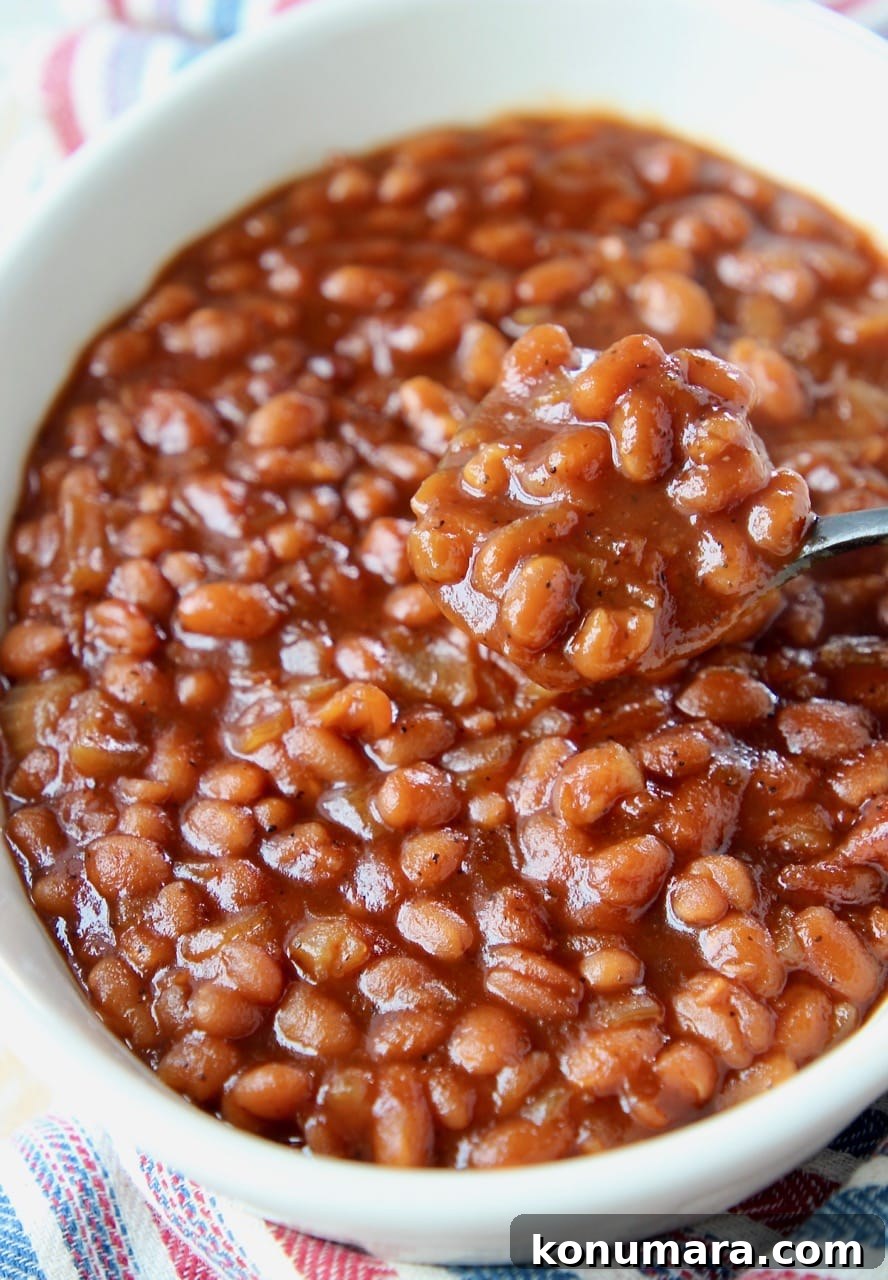Dad's Signature Smoky BBQ Baked Beans Recipe 2 Baked beans in white bowl with black and silver spoon lifting beans out of the bowl