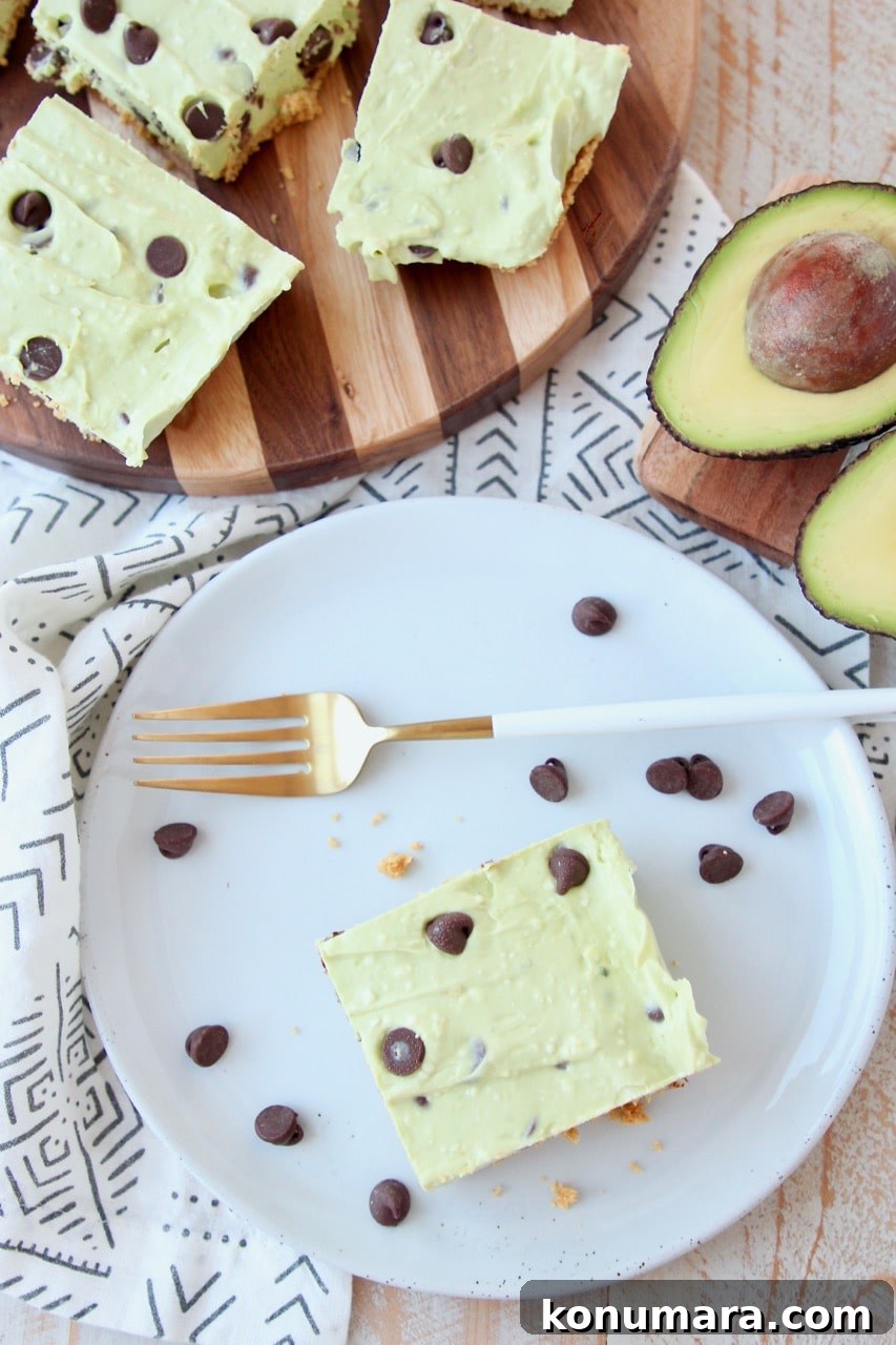 Overhead shot of avocado cheesecake bars on plate and cutting board with chocolate chips and halved avocado