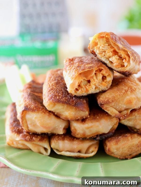 Slow Cooker Buffalo Chicken Recipe 9 Buffalo chicken egg rolls piled up on a green plate with one egg roll cut in half on top, with cheddar cheese and a cheese grater in the background