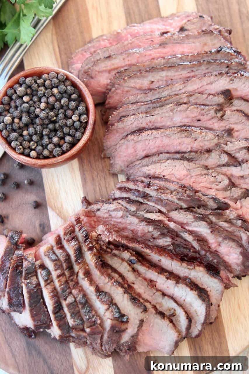 Sliced tri tip steak on wood cutting board with a bowl of black peppercorns
