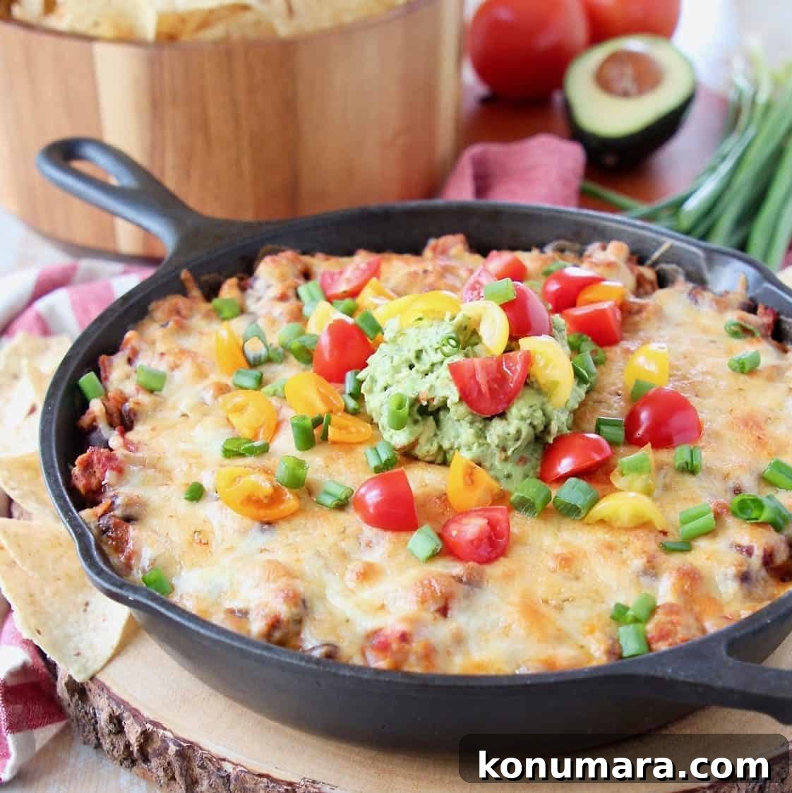 Baked taco dip in a cast iron skillet, sitting on a wood cutting board with a bowl of chips in the background