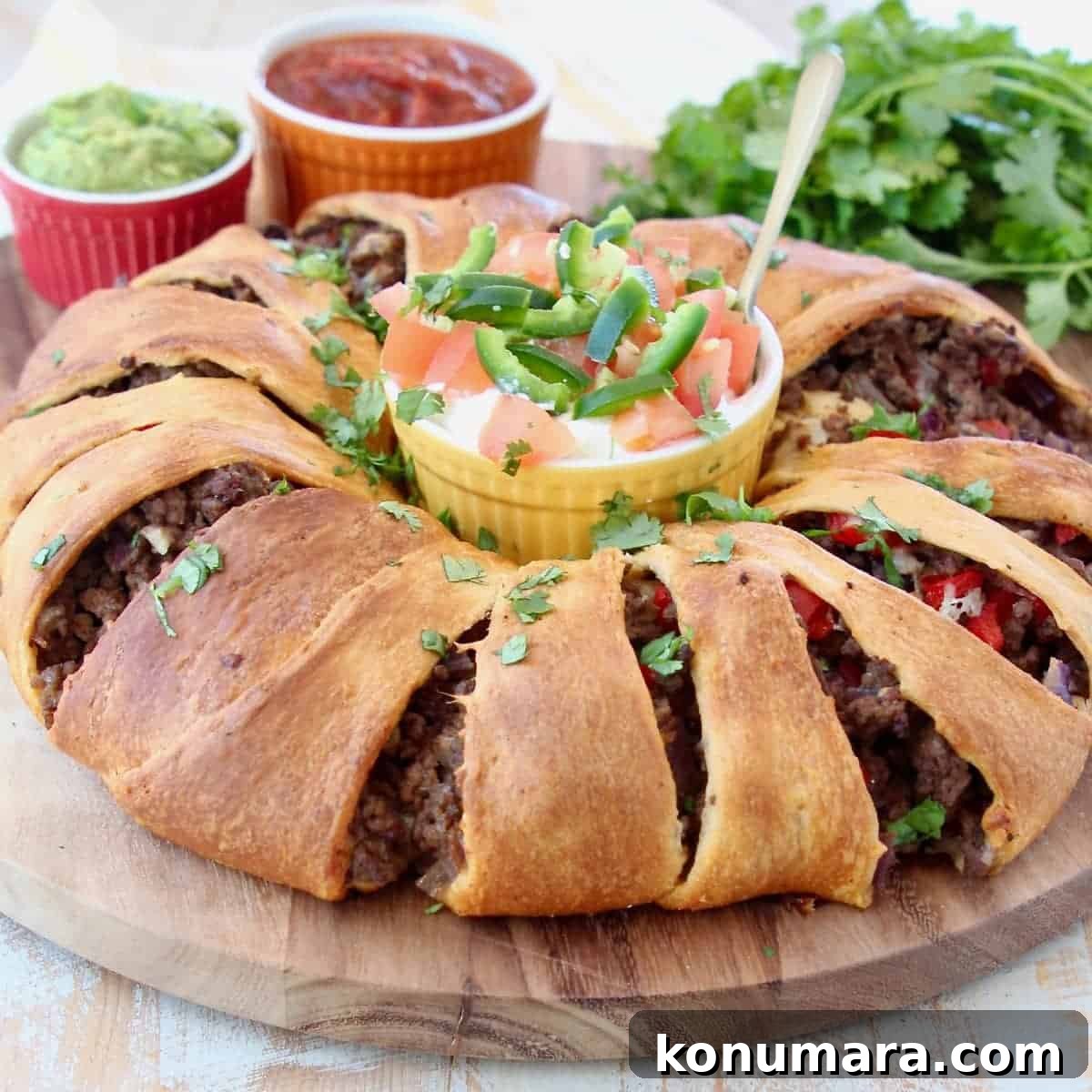 Taco ring with crescent rolls on wood cutting board with ramekins of sour cream, salsa and guacamole