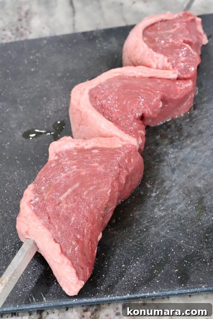 Rotisserie Picanha Recipe: Juicy Brazilian Rump Cap on the Spit 6 three pieces of steak skewered on a rotisserie rod on a cutting board
