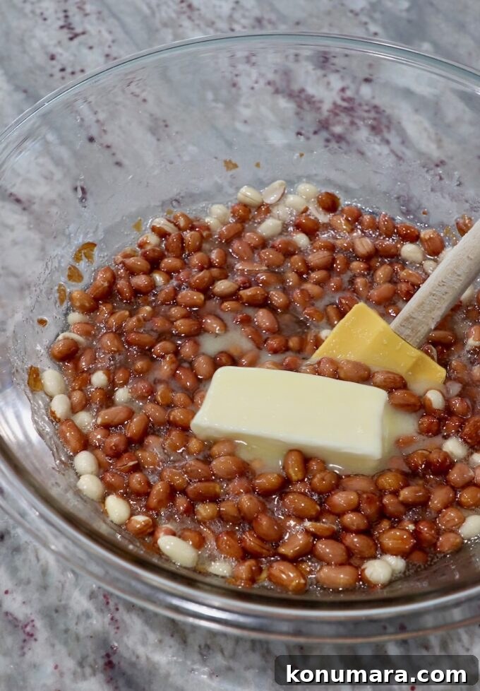 spatula stirring a stick of butter with microwaved sugar, corn syrup and raw peanuts in a large glass bowl