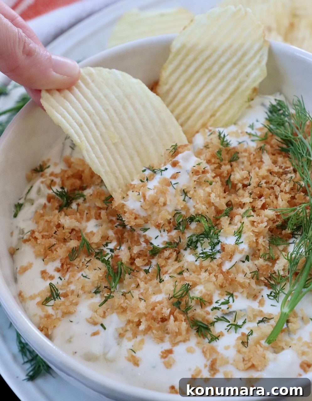 Pickle dip in a bowl topped with toasted breadcrumbs, with a chip dipped into the bowl of dip