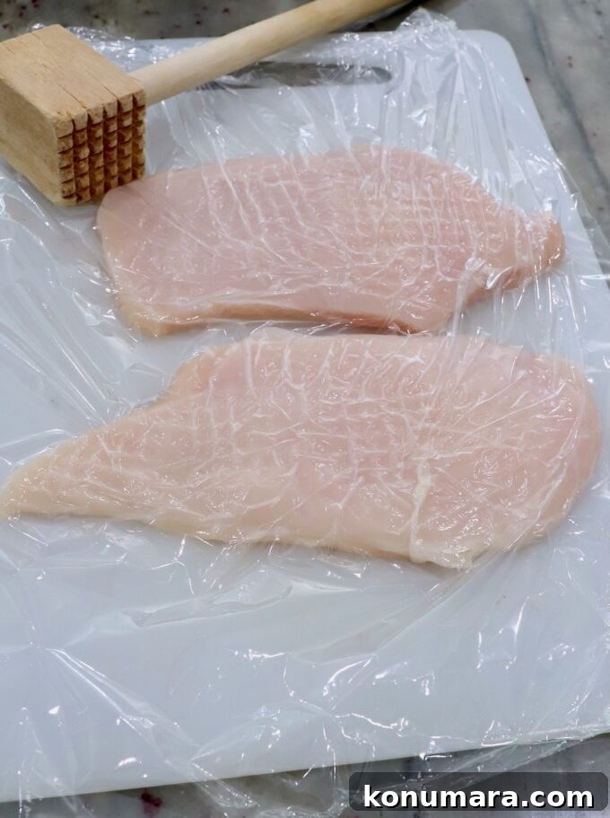 Crispy Parmesan-Crusted Chicken Recipe 4 chicken breasts on cutting board covered with plastic wrap with meat tenderizer on the side