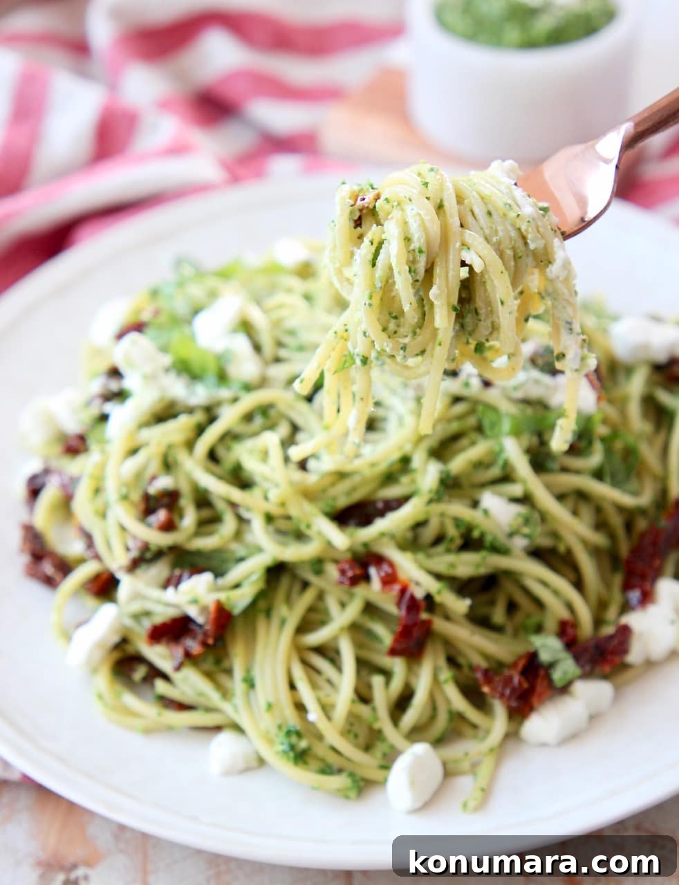 Pesto spaghetti wrapped around a copper fork, held over a plate of pesto spaghetti with crumbled goat cheese and sun dried tomatoes