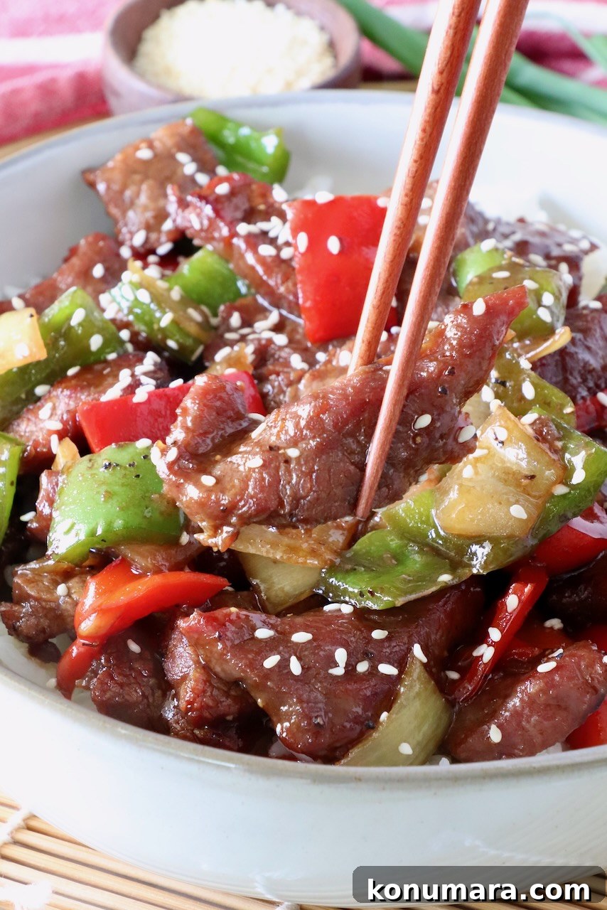 Pepper Steak Stir-Fry Recipe with Bell Peppers 2 chopsticks holding a piece of steak over a bowl of cooked steak and peppers