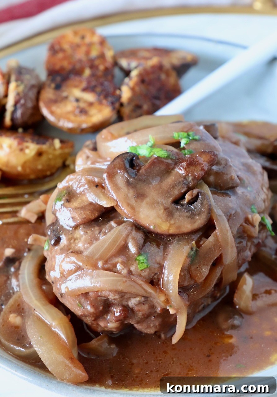 How to Make Chopped Steak with Mushroom Gravy 2 chopped steak on a plate covered with cooked mushrooms and onions, and red wine gravy, with roasted potatoes and a fork on the side of the plate