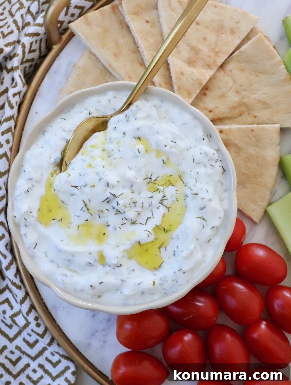 tzatziki sauce in a bowl with a spoon with olive oil drizzled on top and slices of pita bread and cherry tomatoes on the side