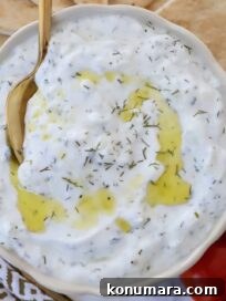 tzatziki sauce in a bowl with a spoon with olive oil drizzled on top and slices of pita bread and cherry tomatoes on the side