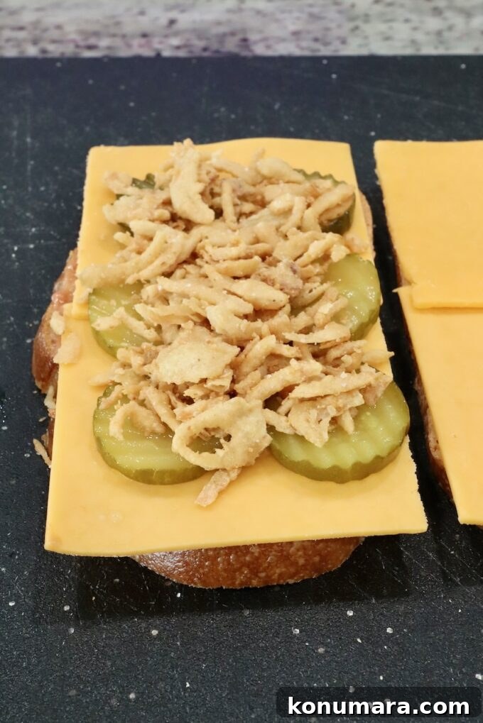 How to Make a Classic Meatloaf Sandwich 6 sliced cheese, pickles, and fried onion strings, on top of sliced bread on a cutting board