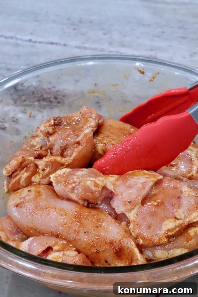 chicken thighs tossed in seasoning in a glass bowl with red kitchen tongs