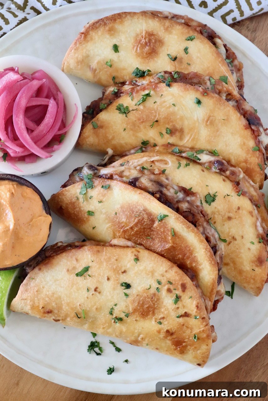 Smoky Black Bean Tacos with Avocado Lime Crema 2 baked tacos stacked up on a plate with a side bowl of pickled onions and a bowl of chipotle sauce