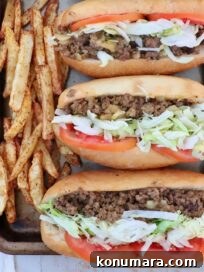 three chopped cheese sandwiches with sliced tomatoes and shredded lettuce lined up on a baking sheet with seasoned, cooked french fries on the side