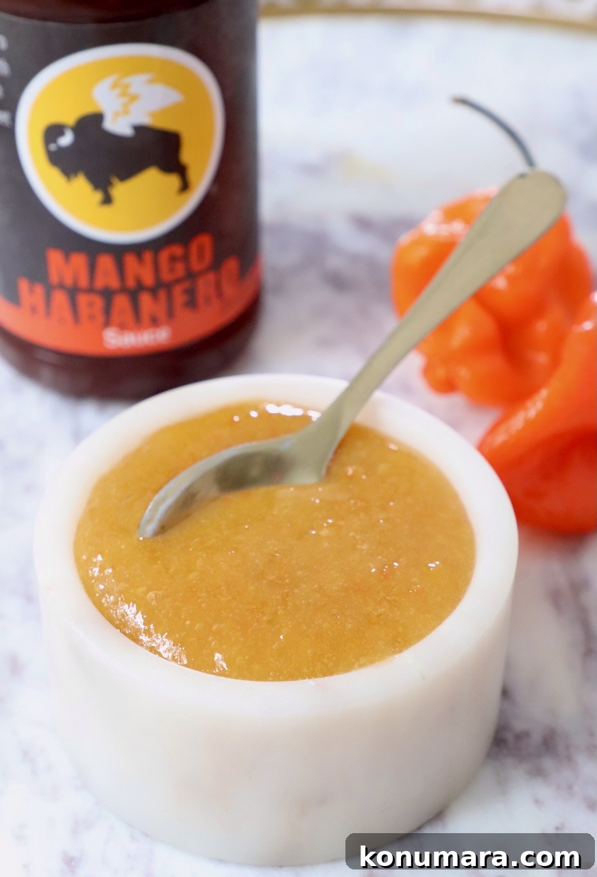 Mango Habanero Sauce Recipe for Wings and Grilled Meats 2 mango habanero sauce in a bowl with a spoon, with two habaneros behind it and a bottle of buffalo wild wings sauce