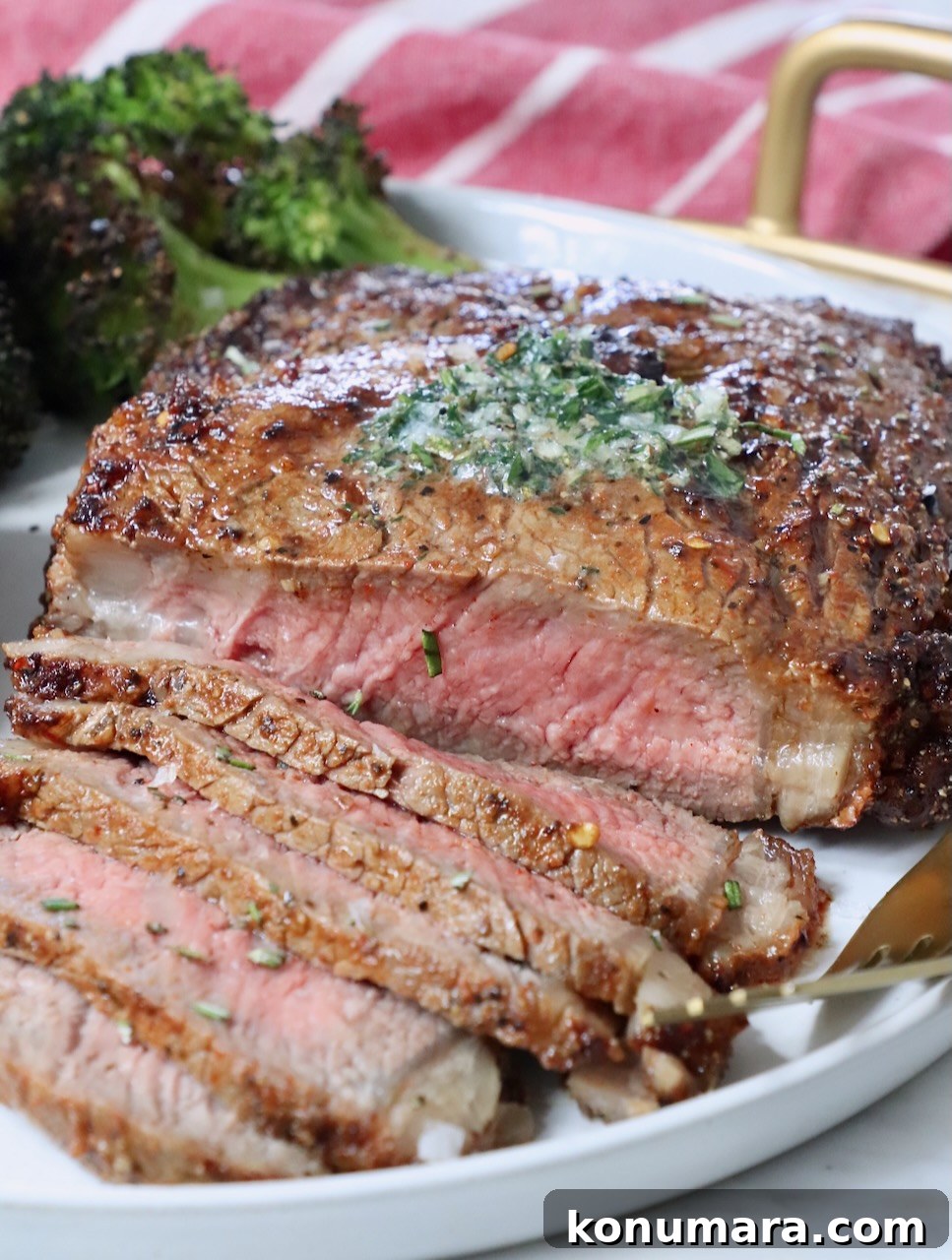 sliced ribeye steak on a plate with herb butter on top of the steak and roasted broccoli in the background on the plate