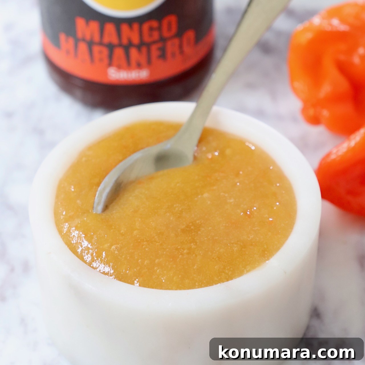 mango habanero sauce in a bowl with a spoon with a bottle of sauce in the background and fresh habanero peppers