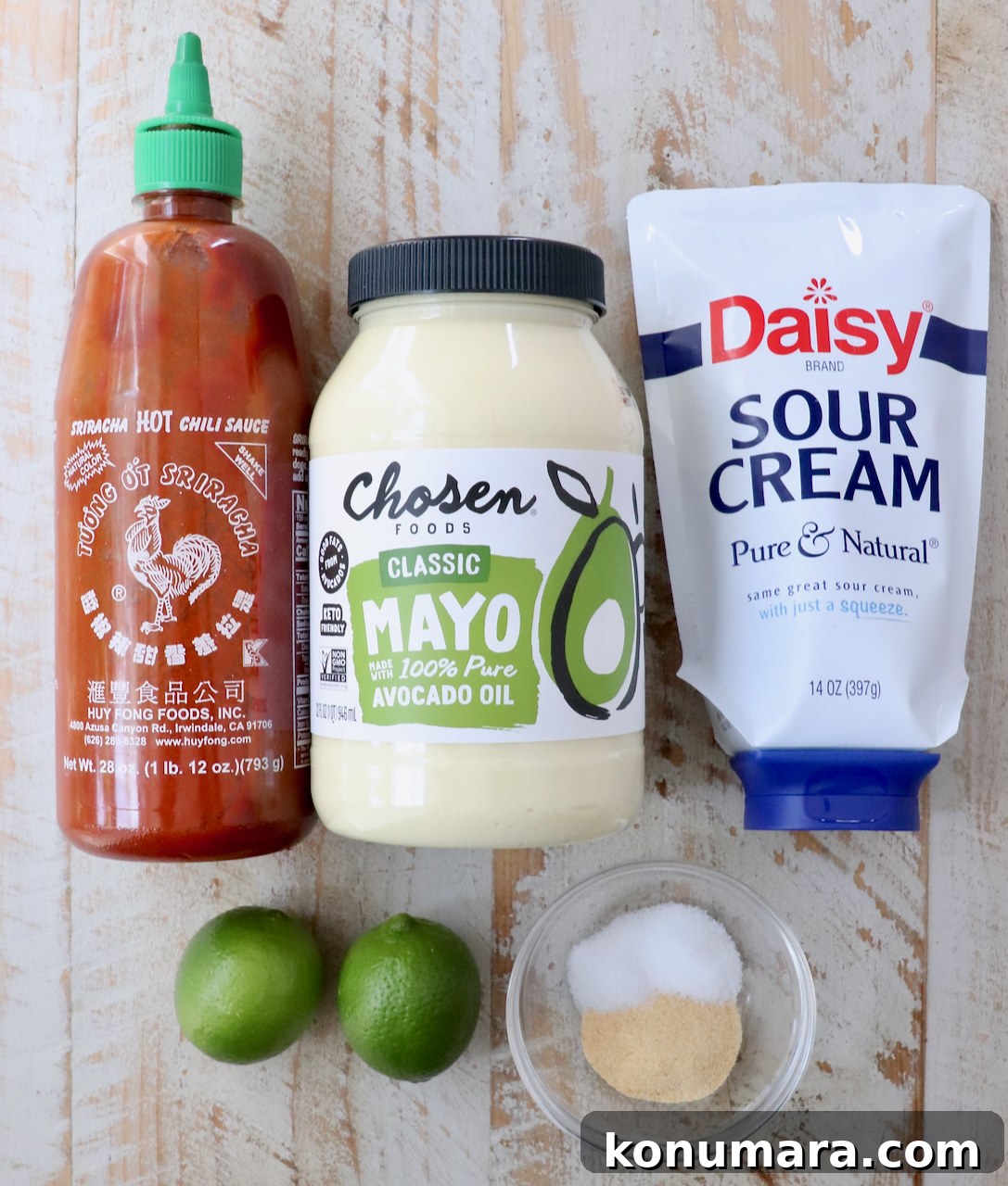 Condiments and ingredients on a wooden surface: a bottle of hot sauce, avocado oil mayo, sour cream, two limes, and a small bowl of salt and garlic powder.