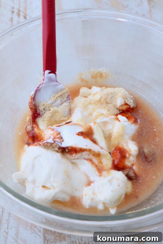 Mixing bowl with white yogurt, hot sauce, and spices, partially mixed with a red spatula.