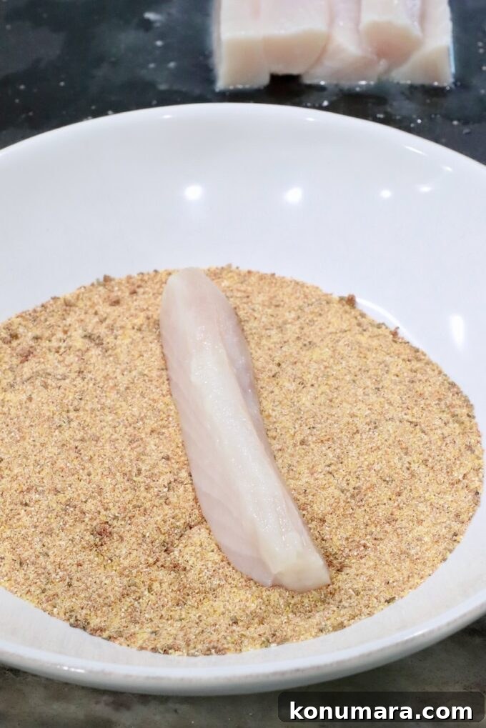 A raw fish fillet sits on seasoned cornmeal in a white bowl, ready for breading. In the background, additional fish fillets are visible on a dark surface.