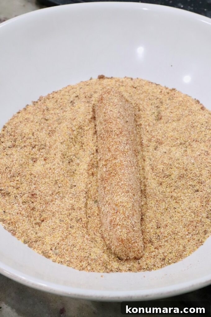 A breaded strip of fish lies on a bed of seasoned cornmeal in a white bowl.