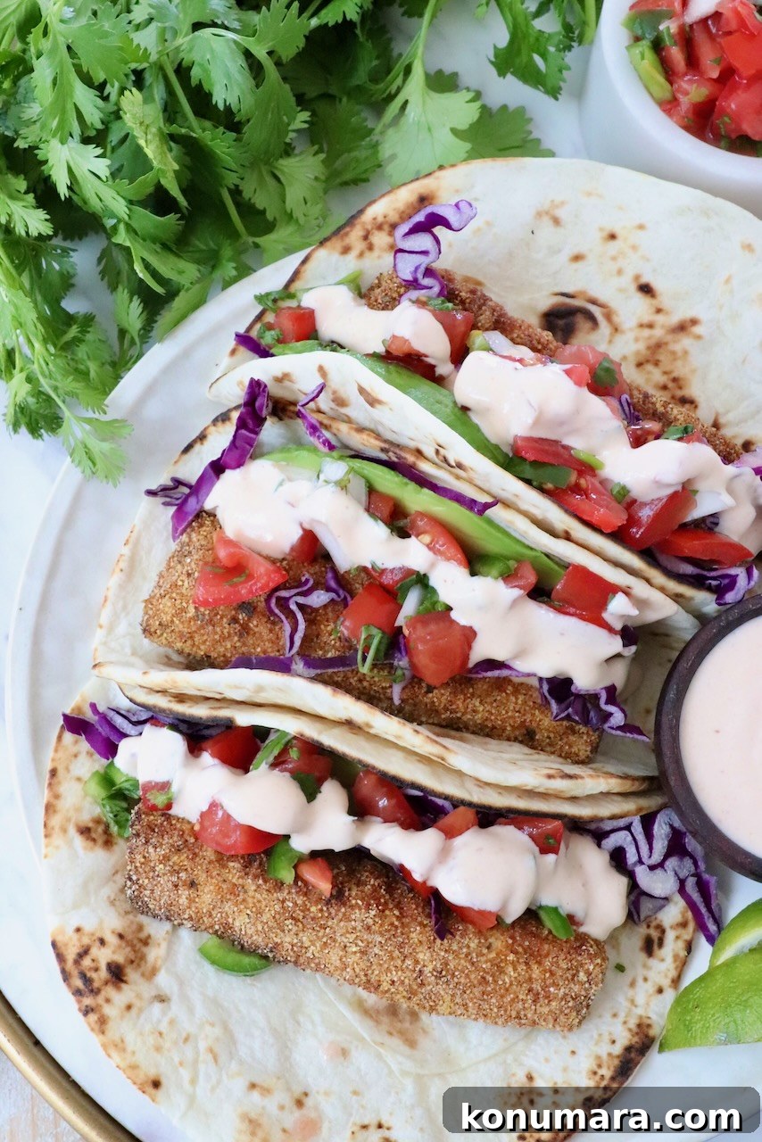 Three tacos with crispy fish, purple cabbage, diced tomatoes, and creamy sauce on a tortilla. They're garnished with fresh cilantro.
