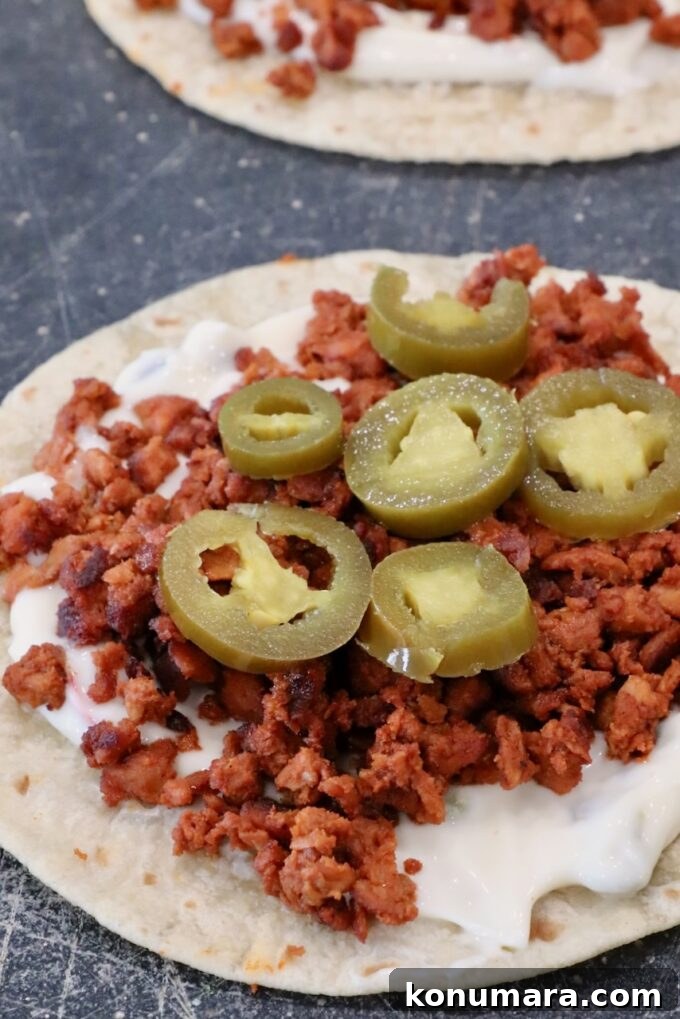 Crispy Chorizo Tacos with Melted Queso 7 Close-up of a tortilla topped with creamy queso dip, crumbled chorizo, and sliced jalapeños on a dark surface