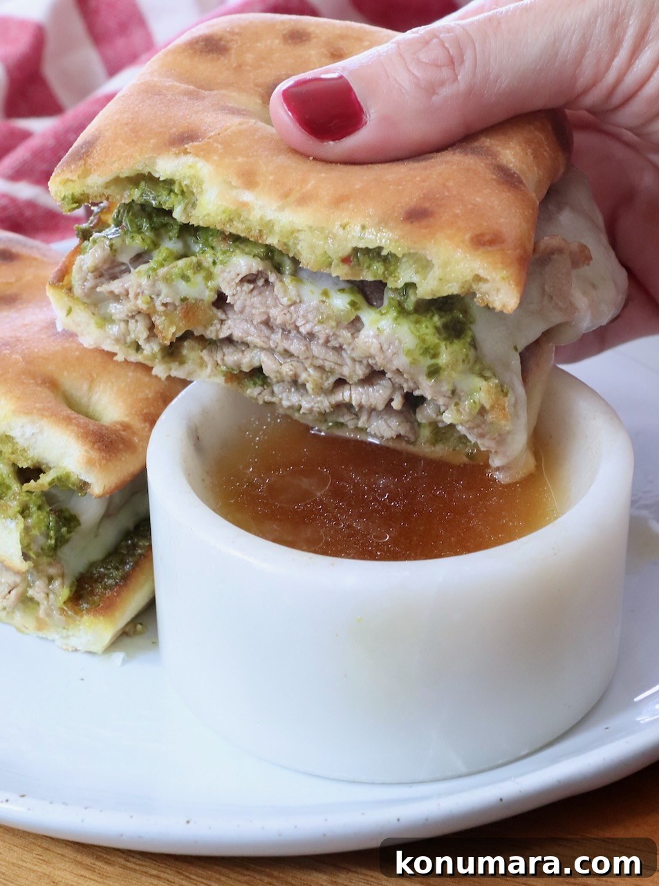 A hand with red nail polish holds a sliced sandwich filled with green chimichurri sauce and cooked steak above a small white bowl of beef broth on a plate.