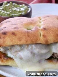 A toasted sandwich with melted cheese and meat is on a white plate. A small bowl of green chimichurri sauce is in the background, with a red cloth nearby.