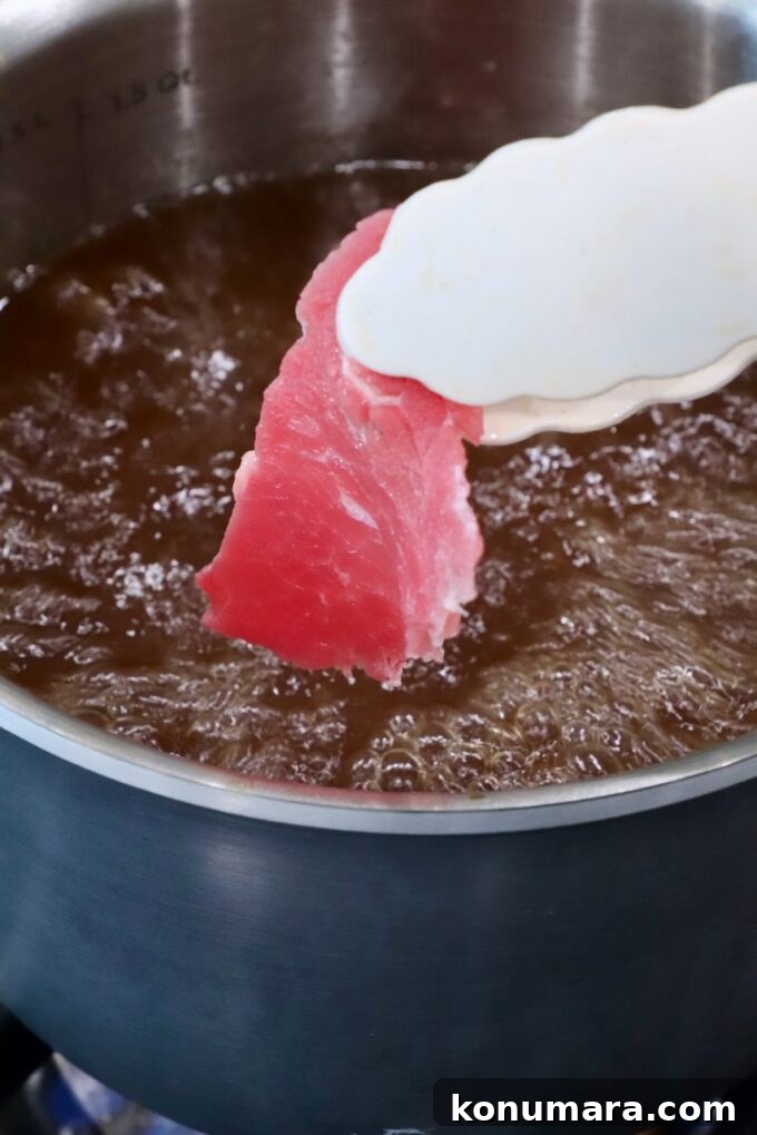 A slice of raw red meat held by tongs is about to be dipped into a pot of bubbling brown broth
