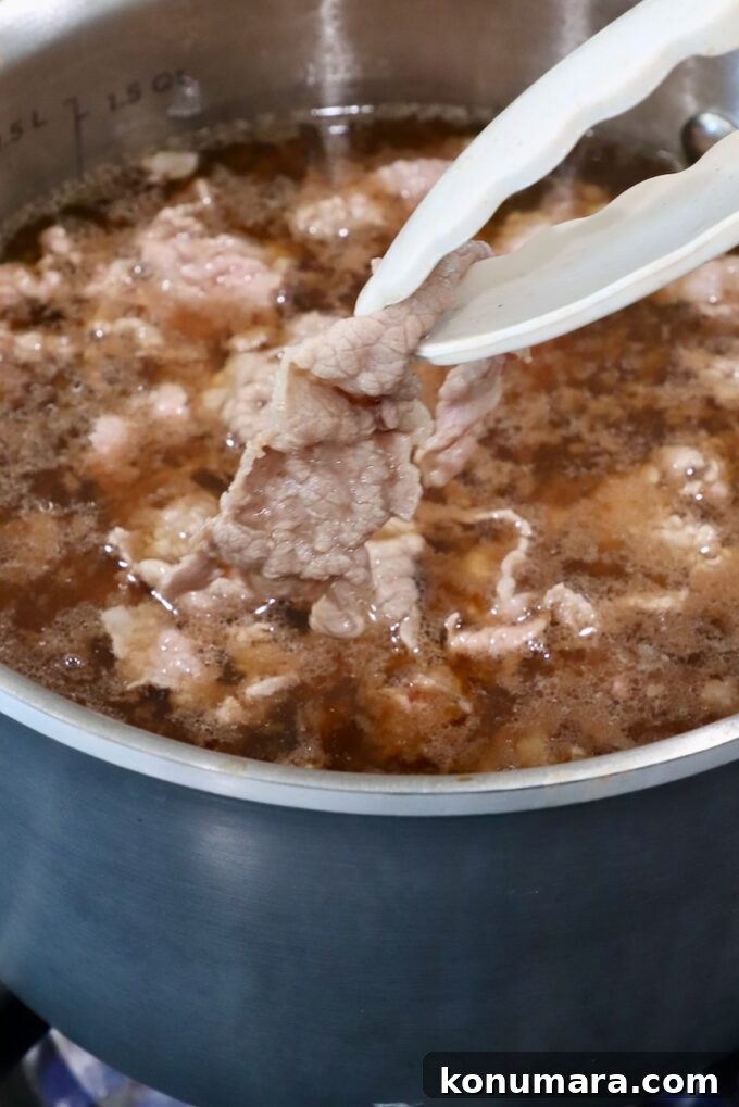 A pot of brown broth with simmering beef slices is being cooked. A pair of tongs holds a piece of cooked beef above the liquid