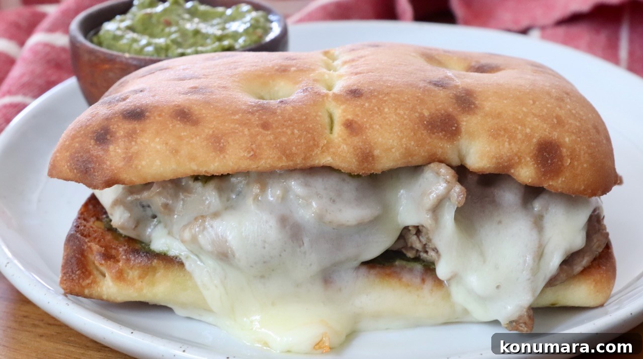 A toasted sandwich with melted cheese and meat is on a white plate. A small bowl of green chimichurri sauce is in the background, with a red cloth nearby.