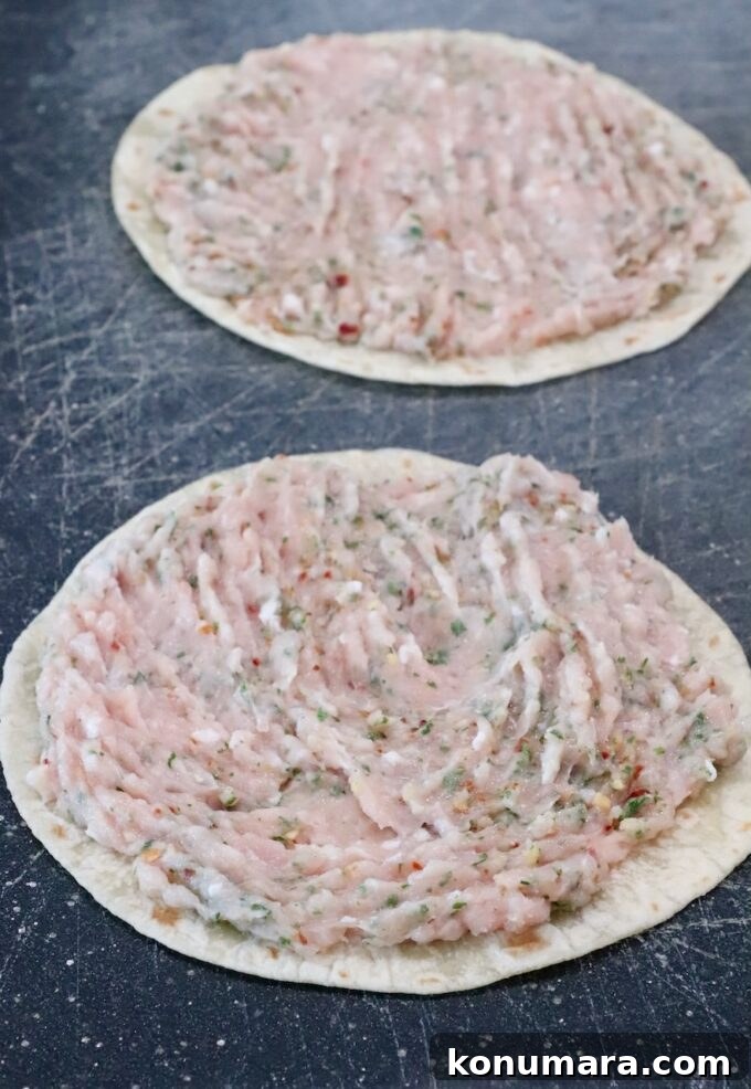 Two tortillas on a dark cutting board, each topped with a spread of raw ground chicken mixed with herbs and spices