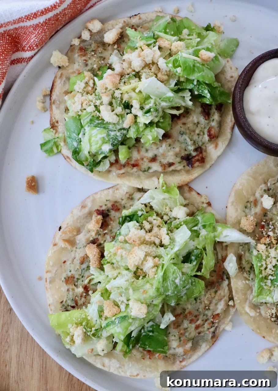 Three mini Caesar salad tacos topped with lettuce, croutons, and creamy dressing on a white plate. A nearby small bowl contains additional dressing.