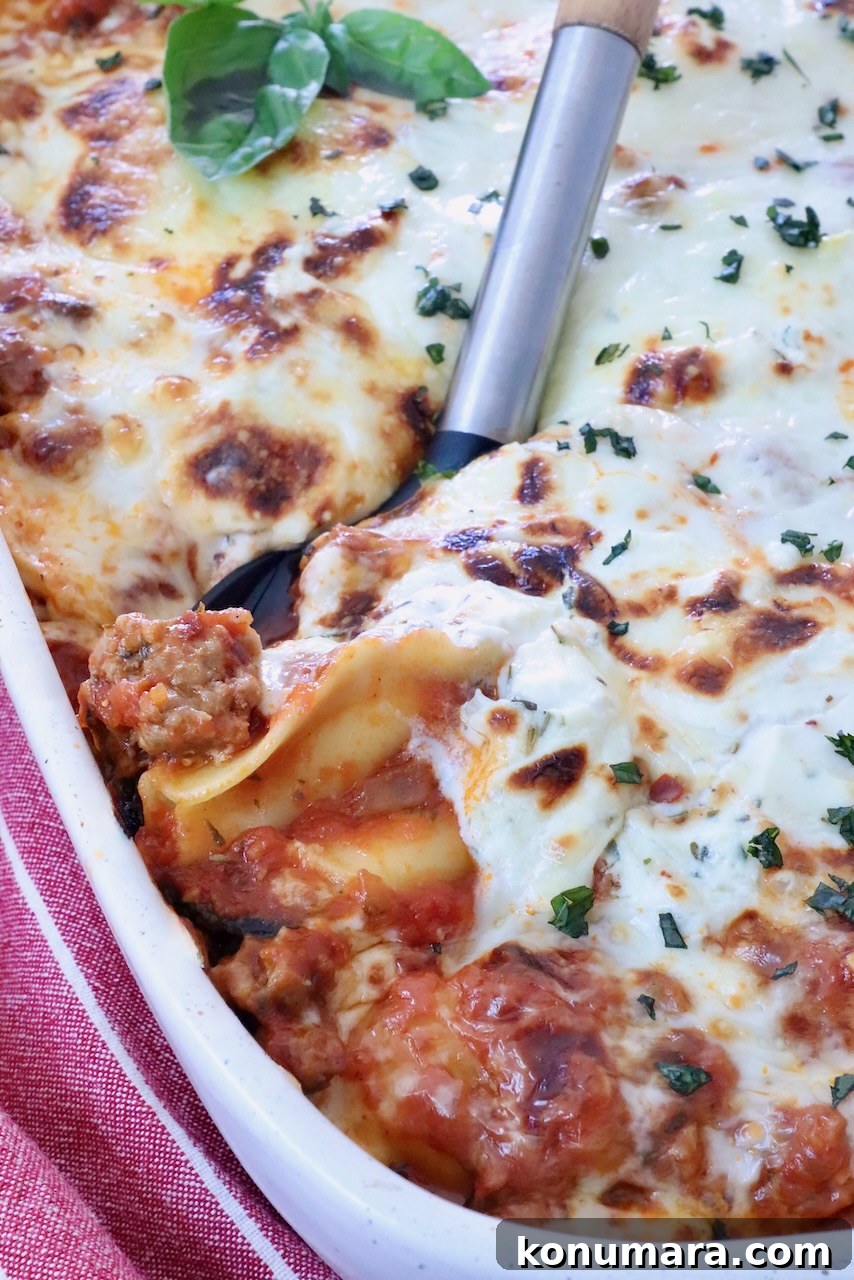 Close-up of a baked ravioli casserole in a white dish, topped with melted cheese and herbs. A serving spatula lifts a portion, showing layers of pasta and meat sauce.