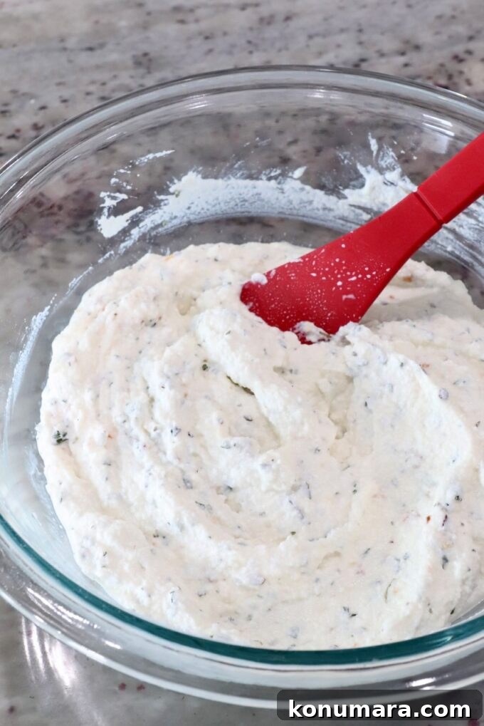 A glass bowl with fluffy, white ricotta cheese mixed with herbs. A red spatula is partially submerged, resting on a smooth marble countertop.