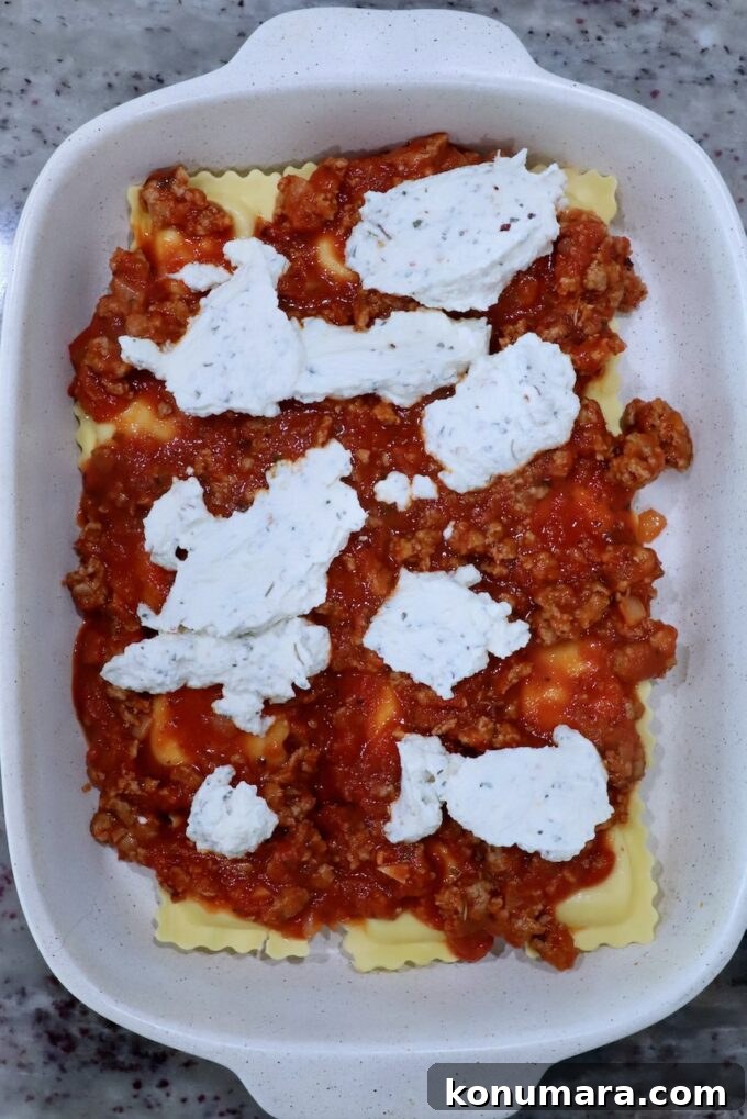 Baked ravioli preparation in a white dish on a speckled countertop. Layers of pasta are topped with meat sauce and dollops of ricotta cheese.