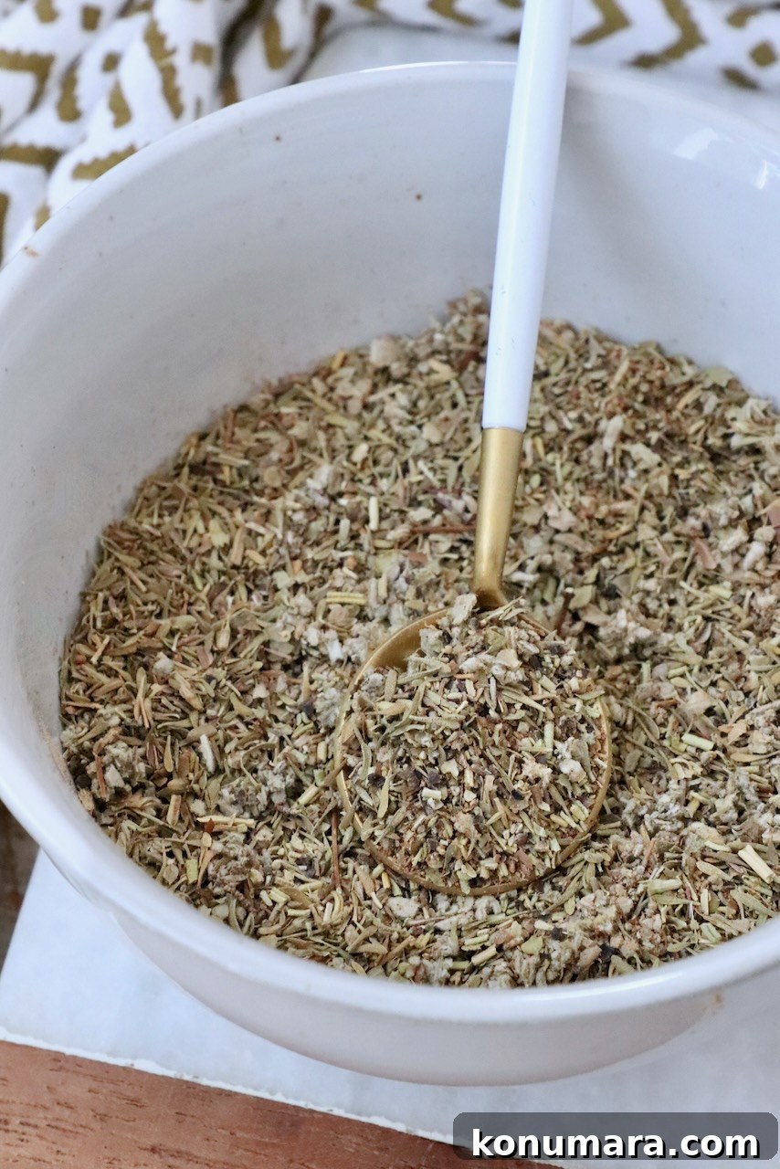 How to Make Poultry Seasoning at Home 2 A white bowl filled with mixed dried herbs and spices, with a gold and white spoon resting inside. A patterned cloth is partially visible in the background.