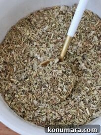 A bowl filled with a coarse, mixed herb blend featuring green and brown hues. A golden spoon with a white handle rests inside.