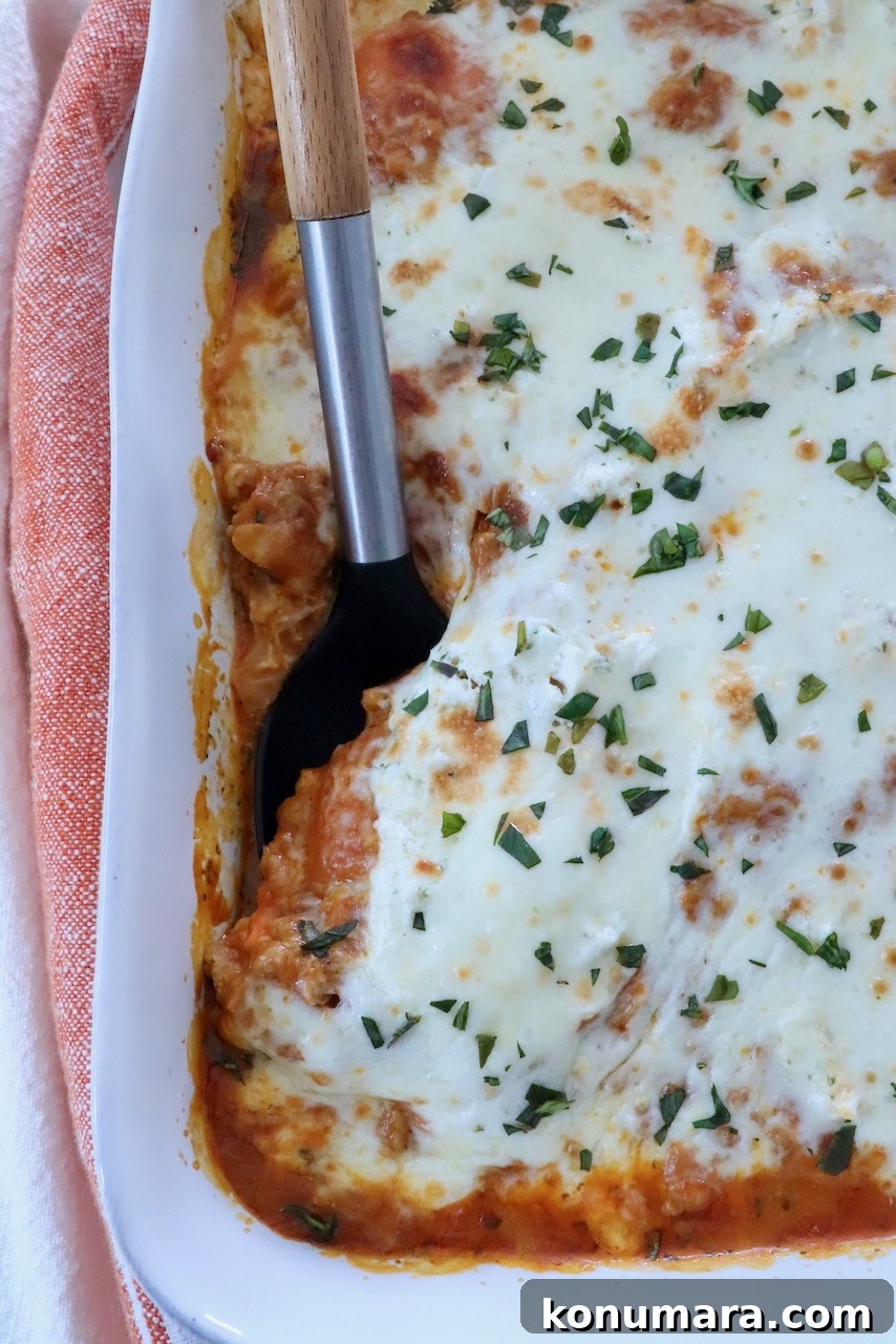 Baked Butternut Squash Ravioli with Brown Butter and Sage 2 Close-up of butternut squash baked ravioli in a white baking dish, featuring melted cheese sprinkled with herbs. A serving has been removed with a black spatula.