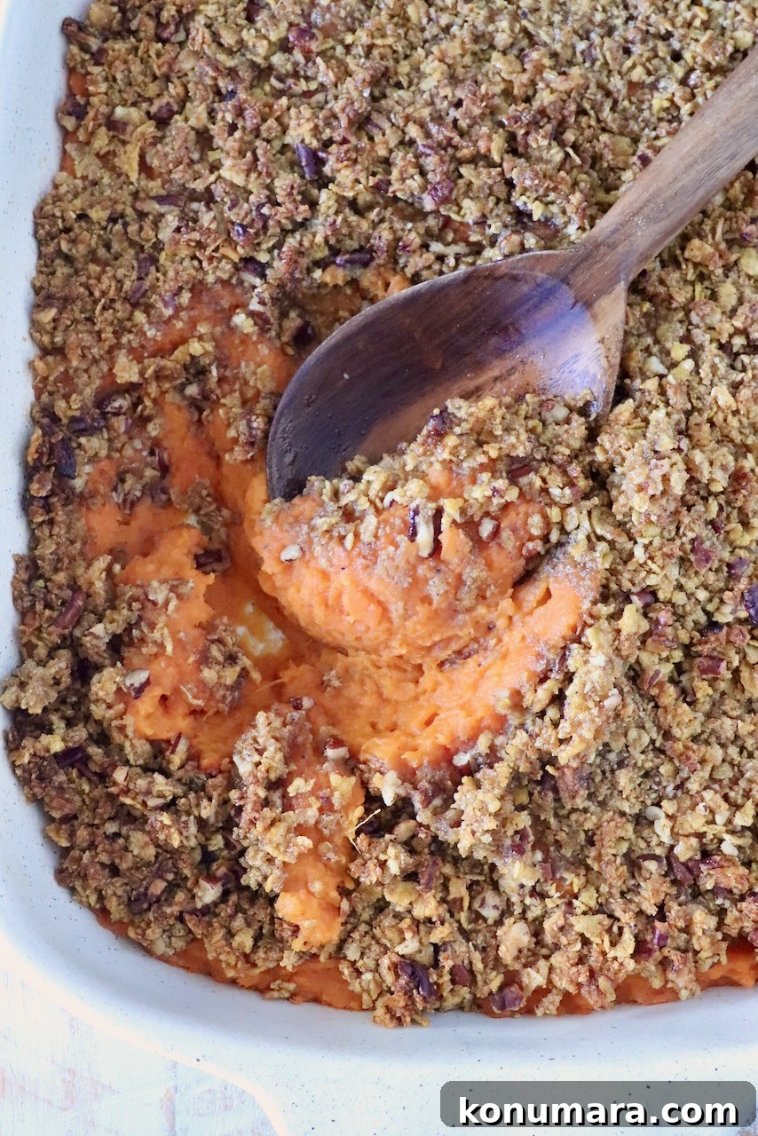 Close-up of a sweet potato casserole with a crunchy pecan topping and a wooden spoon scooping the orange sweet potato filling out from the dish