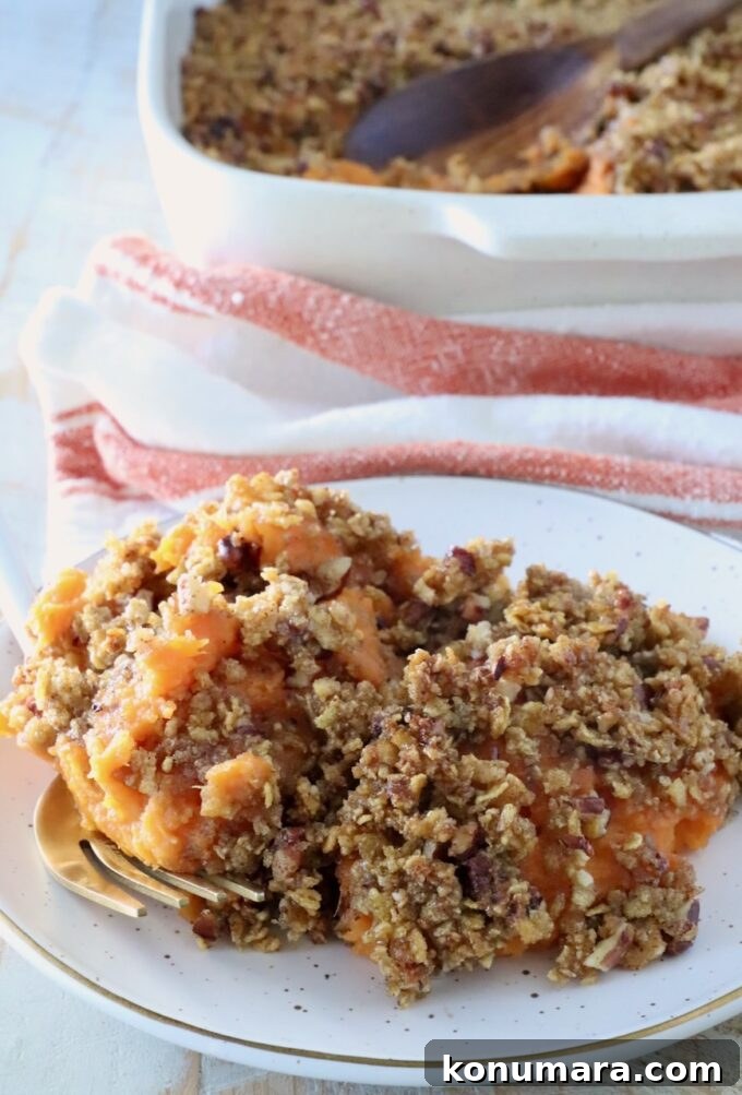 Plate of sweet potato casserole topped with a crispy pecan-crumb layer. A serving spoon lies nearby; a baking dish and towel are in the background.