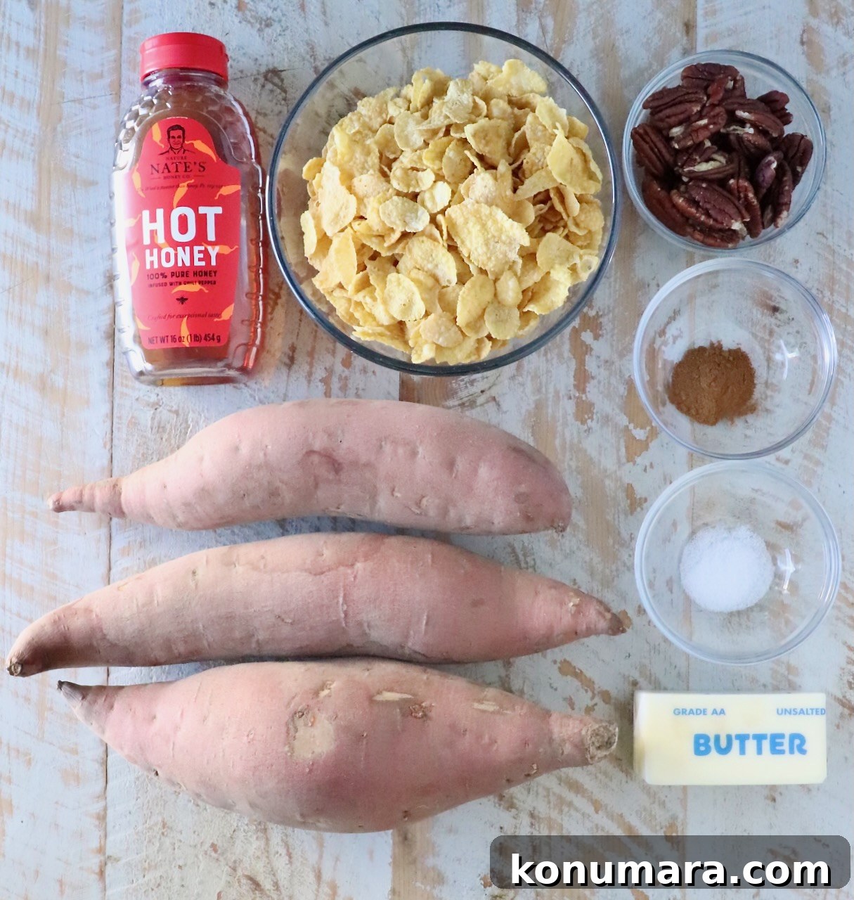 Ingredients for a sweet potato dish on a wooden surface: three raw sweet potatoes, a bottle of hot honey, cornflakes in a bowl, pecans, cinnamon, salt, and a stick of butter.