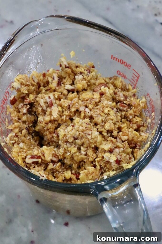 A glass measuring cup filled with a crumbly mix of corn flakes and chopped nuts sits on a speckled granite countertop