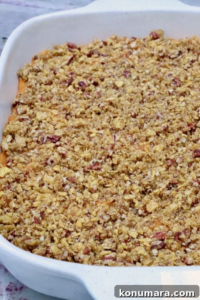 A white ceramic dish filled with a golden-brown, crumbly topping covering a sweet potato casserole, with visible pecan pieces throughout