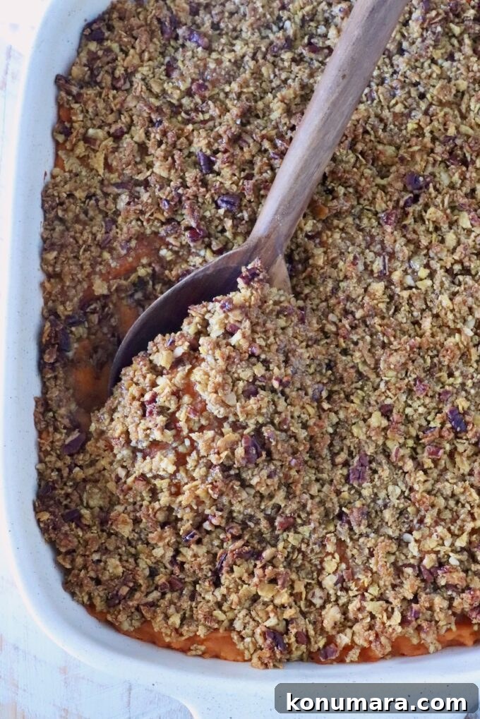 A white baking dish holds a sweet potato casserole topped with a crumbly pecan and corn flake crust. A wooden spoon scoops a portion, revealing orange filling.